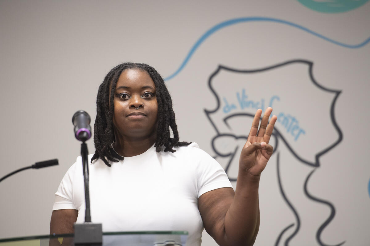 A photo of a woma from the chest up standing. She is holding one hand up with three fingers pointed in the air. She is standing behind a podium with a microphone