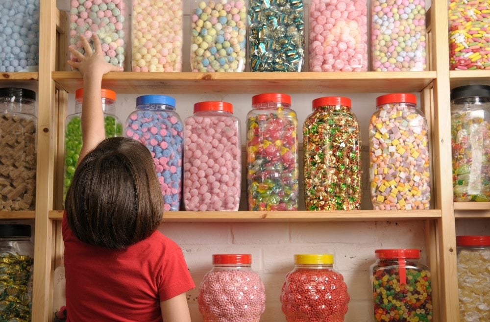 A child stands in a sweet shop gawking at a myriad of choices of brightly colored candies in big glass jars stored in rows on tall shelves in piece about AI marketing tools and the plethora of choices.