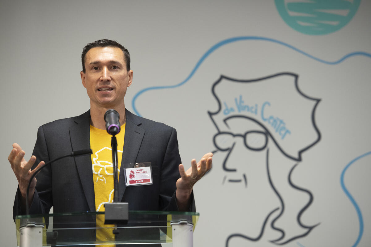 A photo of a man from the wasit up stading behind a podium. He is speaking and gesturing his hands, with his palms facing upwards. 