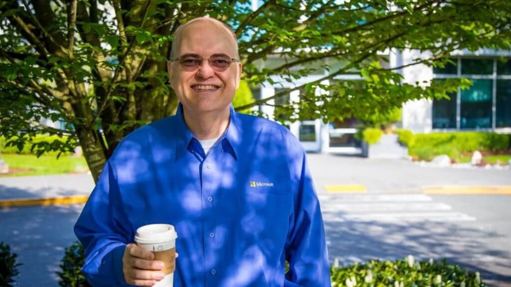 A Microsoft employee smiles as he stands outside of a Microsoft office with a cup of coffee in his hand.