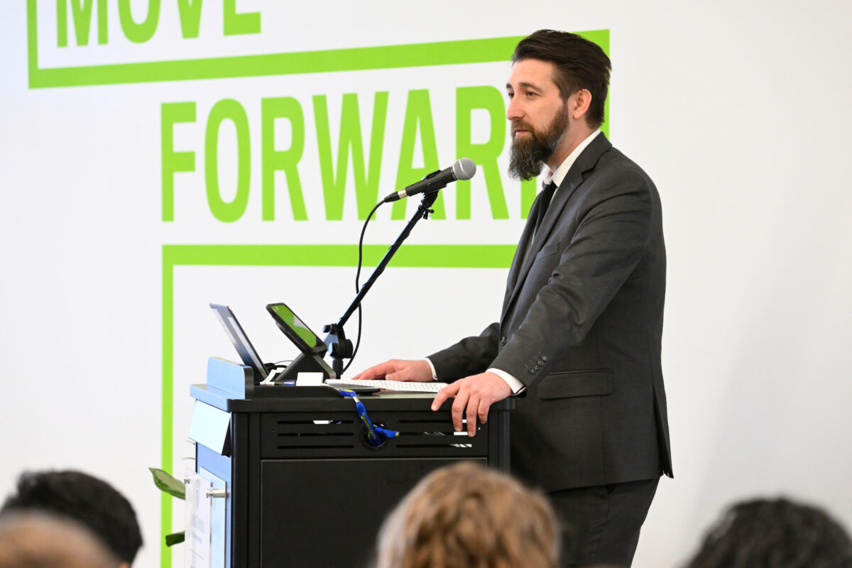 Josh Ambrose in charcoal suit in front of white wall with green text