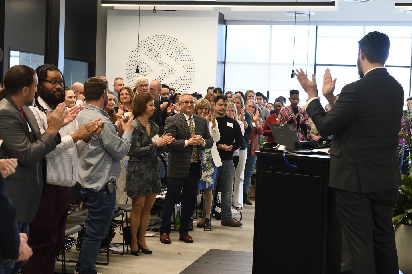 A crowd of people stand up and applaud in JHU's Pava Center. One person is standing at a podium in front of all the others, also clapping.