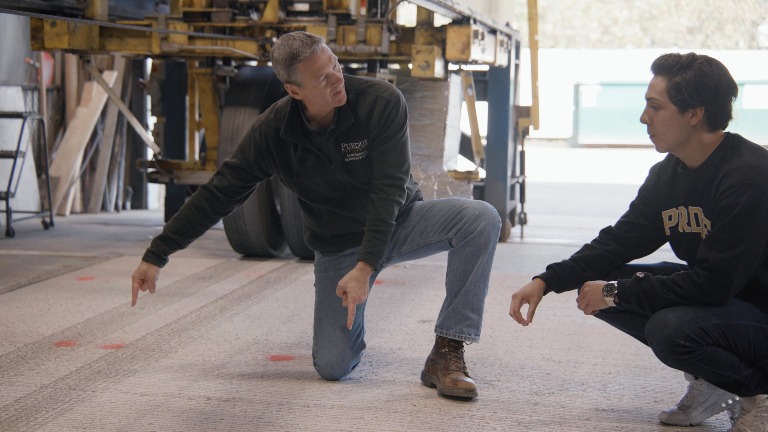 Purdue professor John Haddock and graduate student Oscar Moncada examine a slab of concrete pavement they tested to handle heavy truckloads with wireless power-transfer technology installed below the surface. The machine behind them is designed to imitate those loads by repeatedly passing half a loaded semi-truck axle across the concrete slab. Credit: Courtesy of Consensus Digital Media