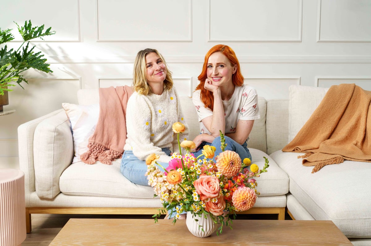 Two women sit on a couch in front of a coffee table holding a large bouquet of flowers