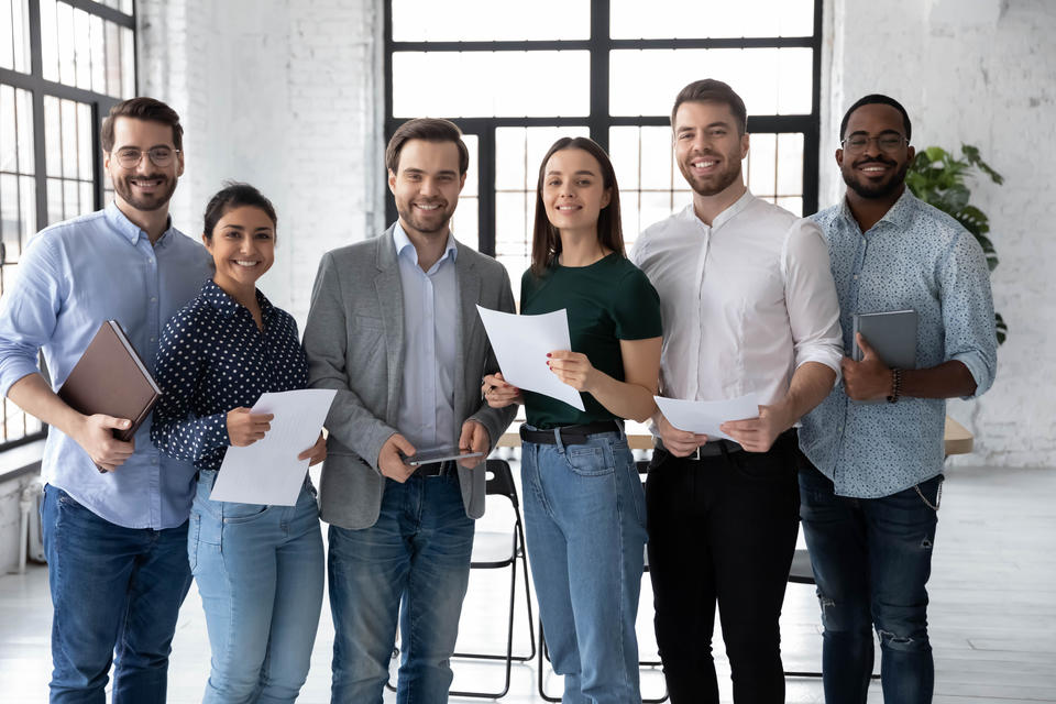 Six smiling, casually dressed young people stand in a line in an office space, holding papers and laptops.