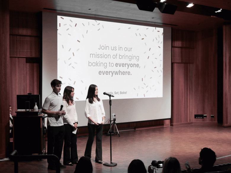 Three students wearing white shirts and black pants stand on stage with their business slogan projected on a screen behind them. (Courtesy photo)