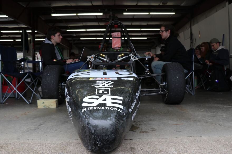 The nose of a racing car is in the foreground with two students at computers on either side. 