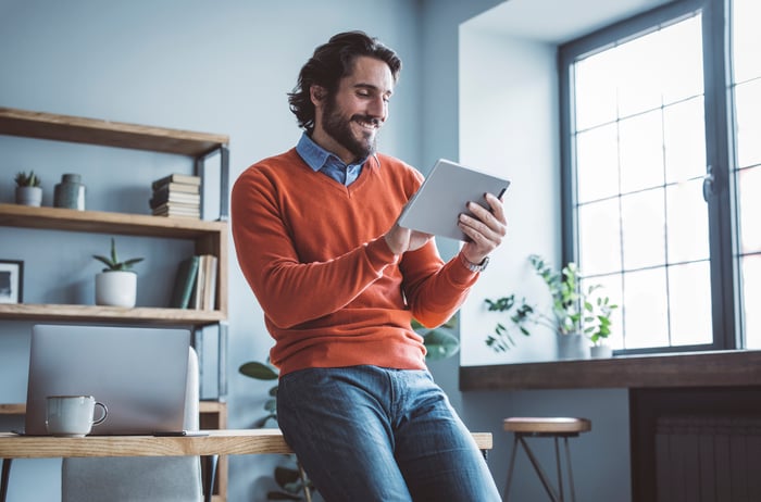 An investor leans against a desk and smiles while looking at something on a tablet.