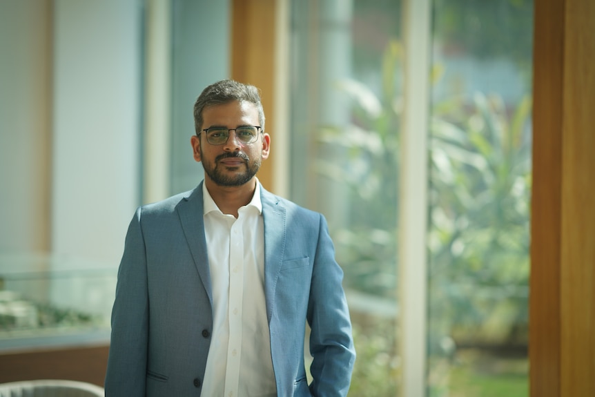 An Indian man dressed in a white shirt and blue blazer smiles in front of see through doors.