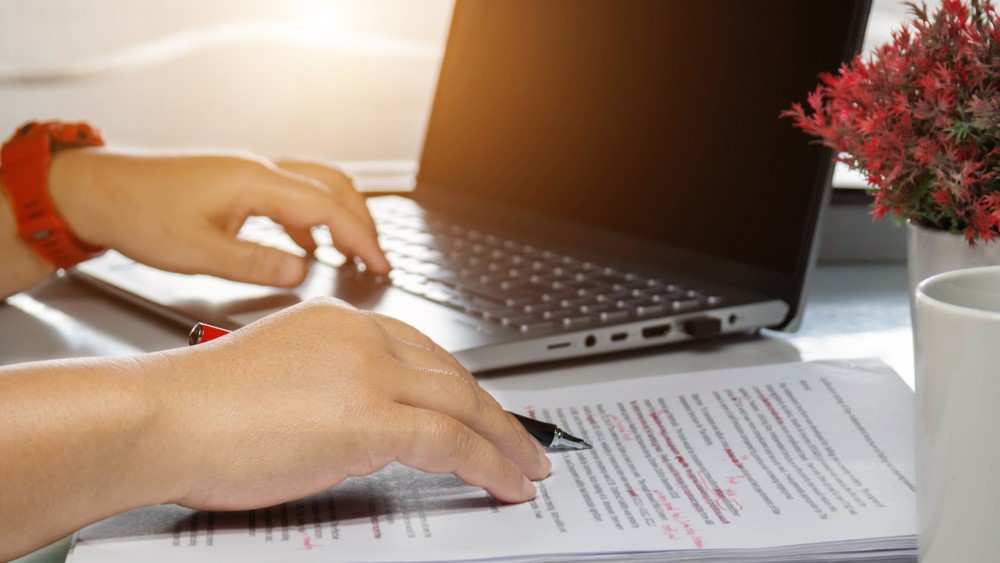 Two human hands write corrections on a piece of paper with a laptop in the background.