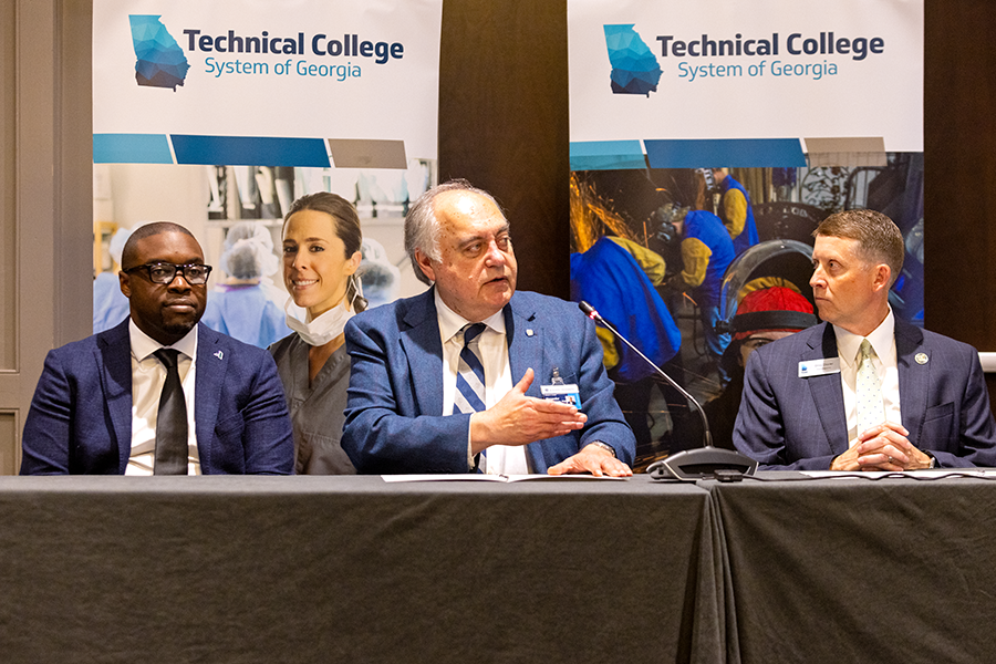 Three men sit at a long table as the man in the middle makes remarks into a microphone.