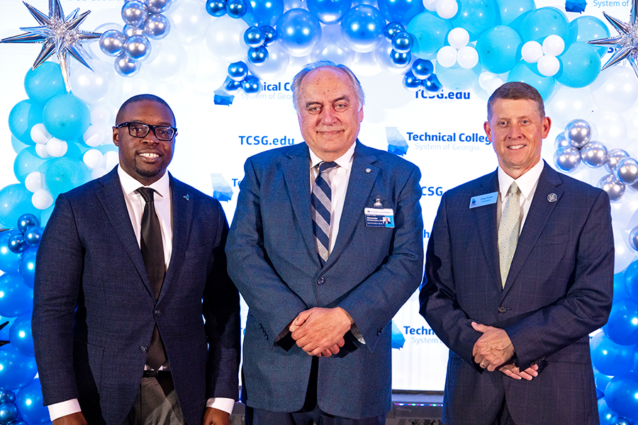 Three men stand in front of a backdrop and pose for a photo following a ceremony.