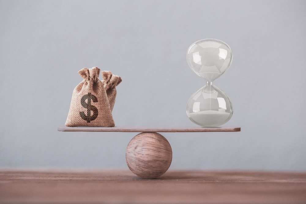 A burlap sack with a dollar sign printed on it and a clear hourglass filled with white sand are balanced on opposite ends of a wooden plank, which is centered on a wooden sphere, depicting the balance between time and money. The background is a neutral gray in piece about vertical AI and its impact on business.