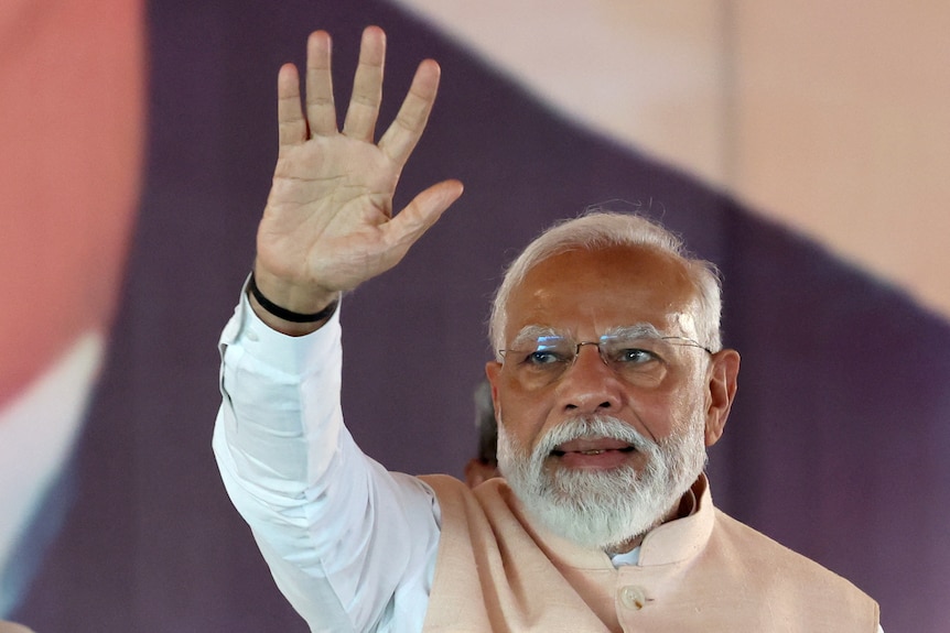 A man with thinning white hair and white beard waives with his right hand