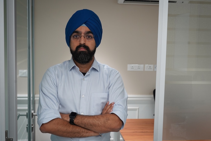 An Indian man with a dark beard and wearing a blue turban stands with his arms crossed.