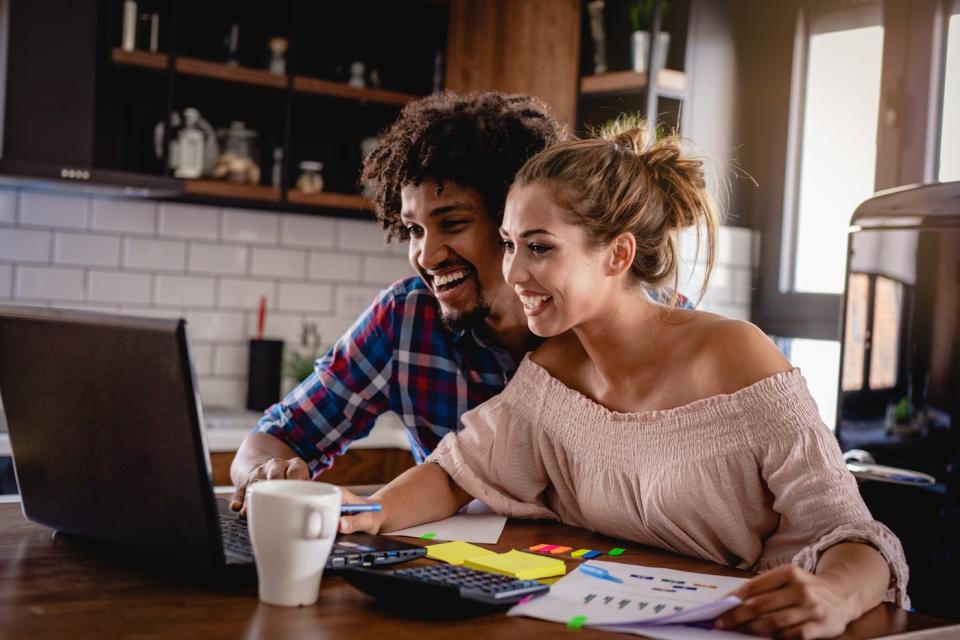 Two investors in a living room smile while looking at something on a laptop.