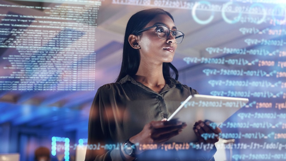 A woman holds a tablet and looks at numbers and symbols on a virtual screen.