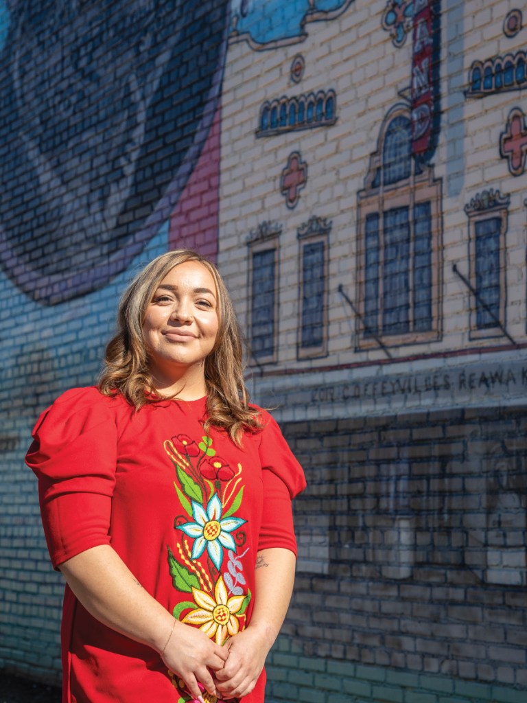 Women in colorful embroidered dress stands in front of mural.