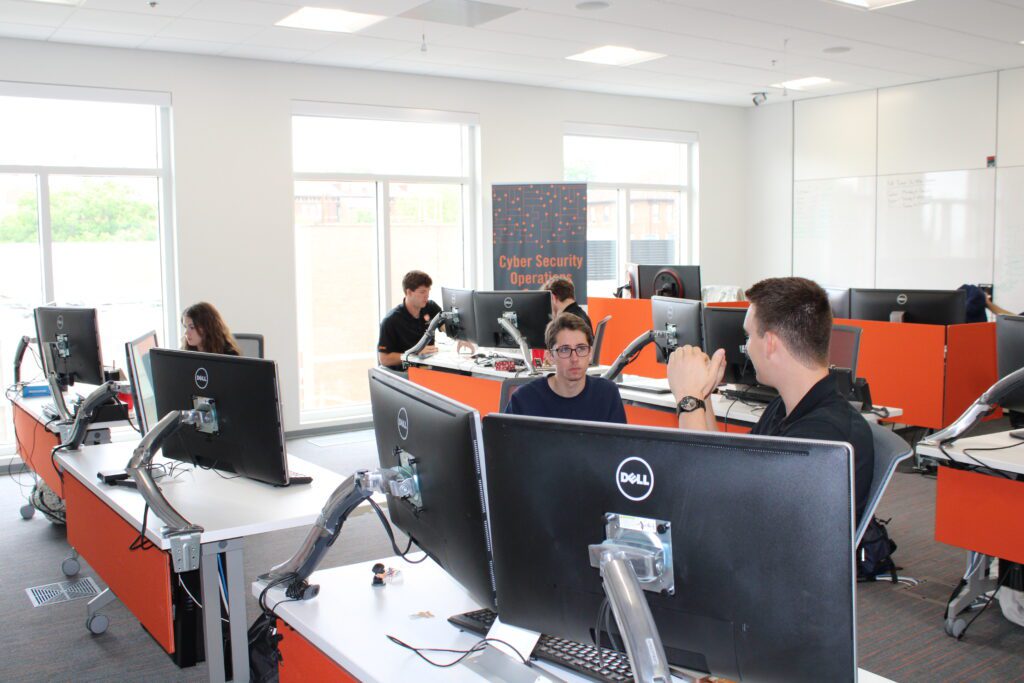 Daniel Harold speaks with another student in the CSOC while sitting in front of computer screens at a desk. 