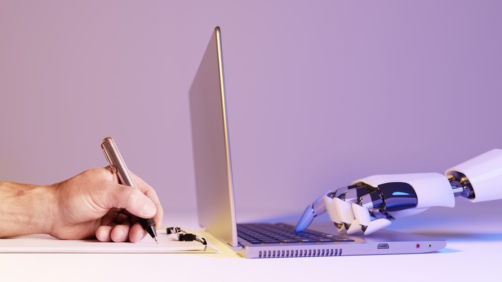 A human hand writing and a robot hand typing on a laptop face each other.