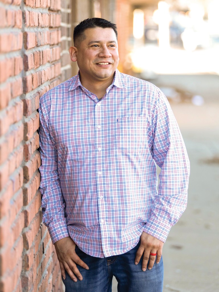 Man in plaid shirt leans up agains brick wall and smiles.