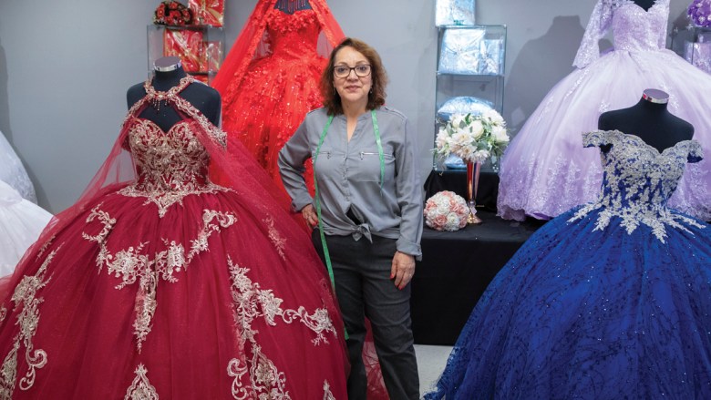 Woman stands amongst numerous mannequins wearing fancy dresses.