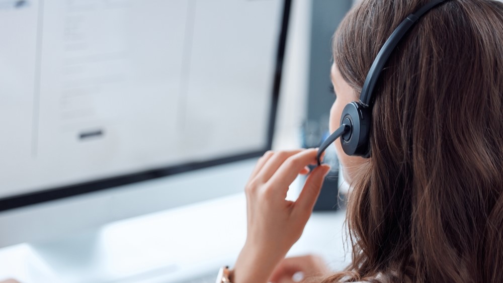 A woman wearing headphones looks at a screen while talking into a mouth piece.