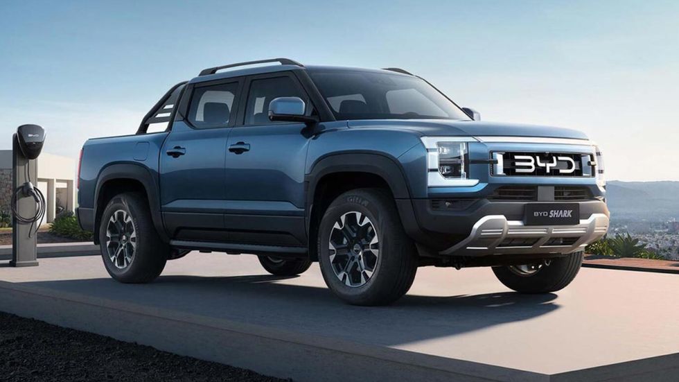 blue four door pickup sits on paved concrete surface with a charging station behind it and with mountains in the background