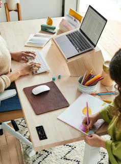 woman works on laptop while a child is drawing on the same table nearby