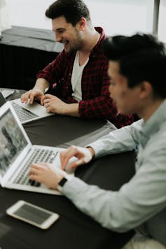 Two men work on laptops, one smiles