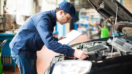 Mechanic working under car bonnet with clipboard in garage