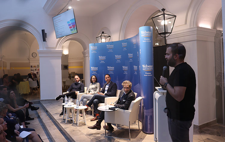 Markus Lang holds a microphone and stands in front of a podium while speaking to a crowd of attendees in the atrium of the Palais Wenkheim. 