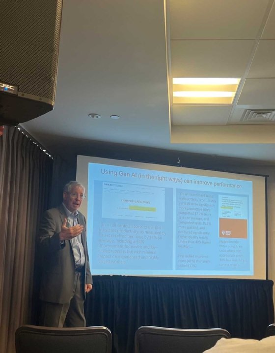 A man stands at the front of a room giving a presentation. The slide behind him reads, 'Using gen AI in the right way can improve performance.'