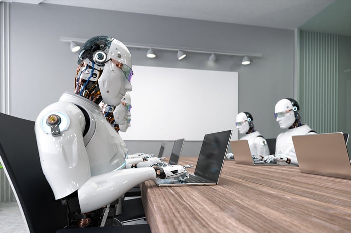 Multiple robots typing on laptops while seated at a conference room table. 