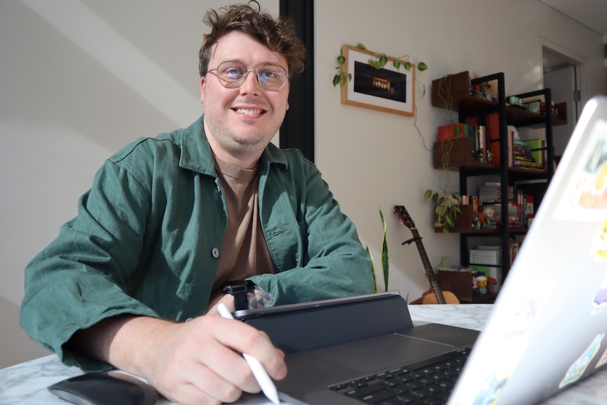 A man with dark, curly hair, wearing a green shirt, sitting at a table with a laptop.