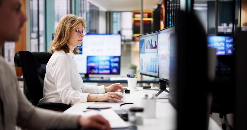 A woman is seated at a desk in a modern office, intently working on two large computer monitors displaying various graphs and data charts. She is wearing glasses and a white shirt. In the background, the office is filled with other desks and computer screens, indicating a busy work environment focused on data analysis in piece about predictive analytics.