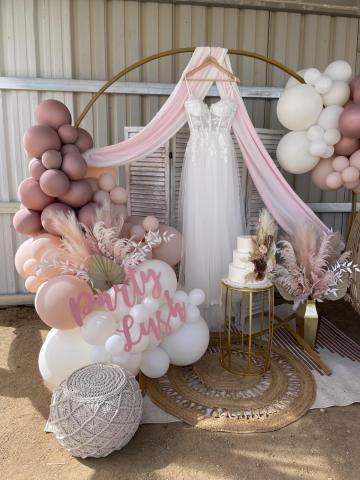 A wedding dress hangs on a wooden hanger from a massive hoop. Balloons, a fake wedding cake, a vase with feathers, a pouf, a small woven rug and a pink wooden cutout that reads “Party Lush” are on display.