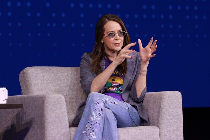 CISA Director Jen Easterly speaks to a crowd at a conference. Woman in blazer, purple-tinted sunglasses, and embroidered jeans, speaks to the audience, sits on an arm chair.