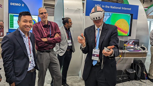 At the DOE booth, two people watch a third who wears VR goggles and gestures with handheld controllers