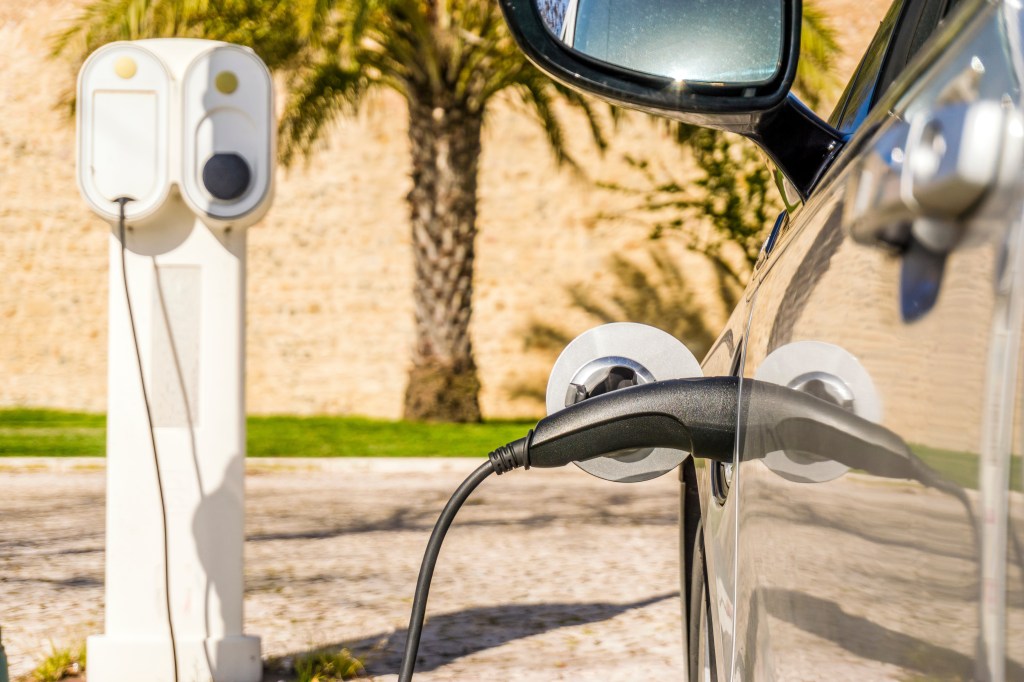 Electric car plugged by cable to charging station with palm tree and old walls as background