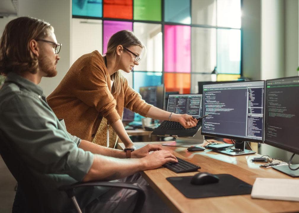 A man working with multiple monitors are engaged in a discussion with his female coworker who points at one of the screens.