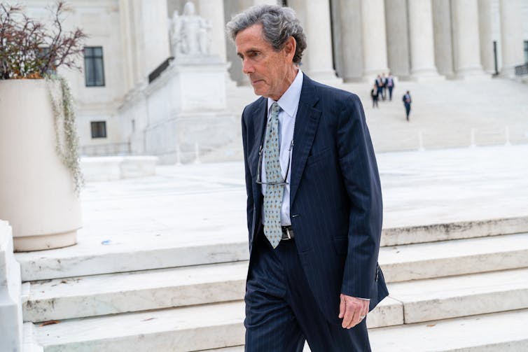 Man in suit walks in front of a large building with white marble columns.